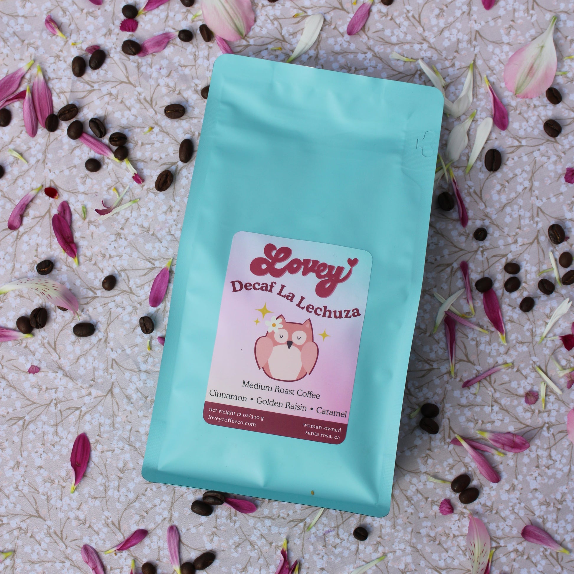 A stand-up coffee bag in light aqua blue with a pink label reading 'Lovey Decaf La Lechuza,' placed on a pale patterned surface scattered with pink flower petals and dark coffee beans.