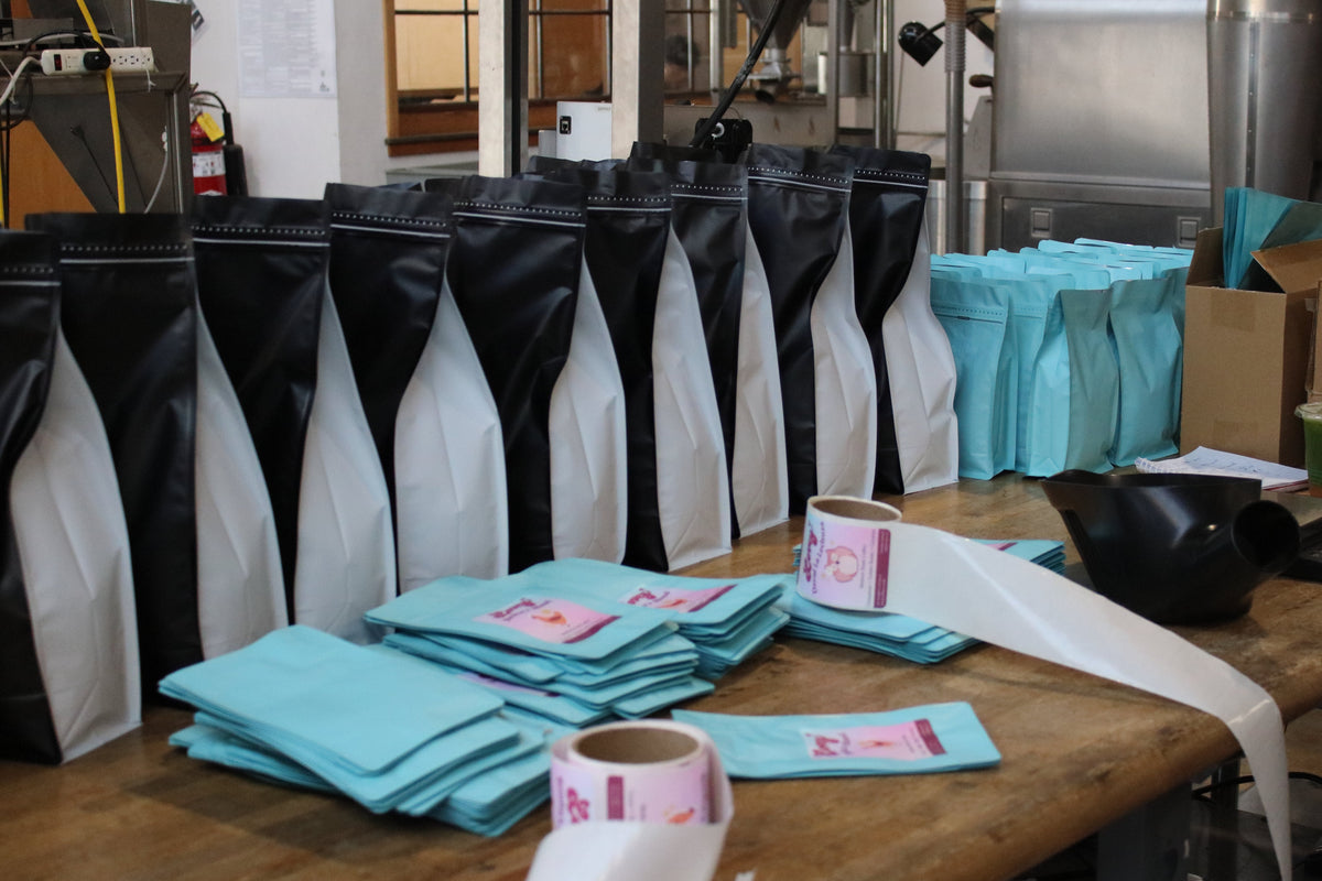 A production line at a coffee roastery showing black and white coffee bags, light blue bags, and rolls of labels on a wooden table.